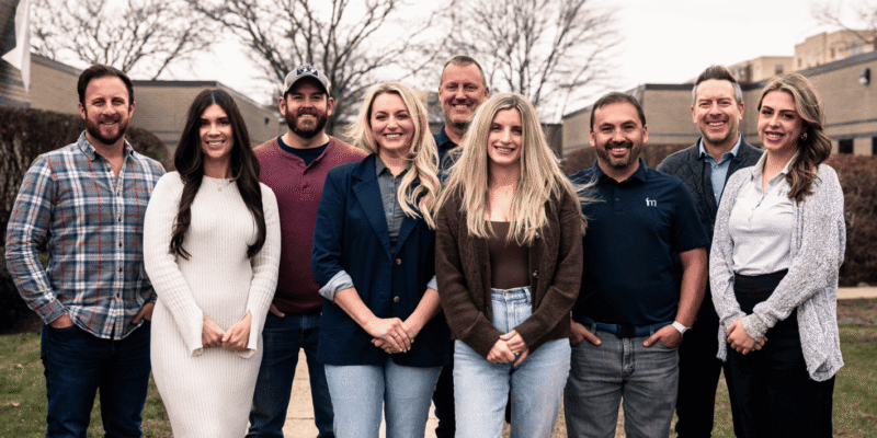 FM team smiles outdoors, dressed casually, with trees and buildings.