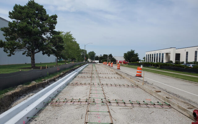 Road construction with exposed rebar, gravel, and orange barrels. Buildings and trees line road.