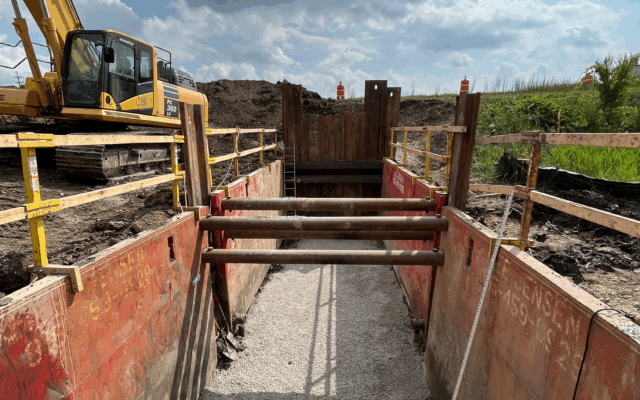 Trench with metal supports, gravel base, excavator in background.