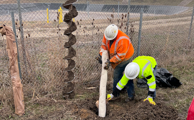 Two workers in safety gear insert cylinder into ground.