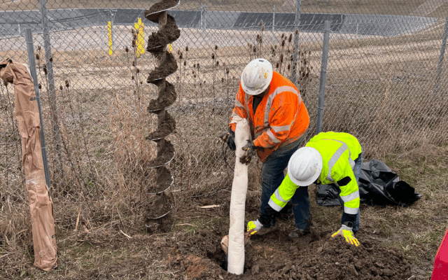 Two workers install cylinder in ground near fence, equipment nearby.