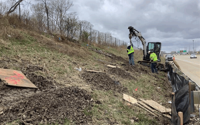 Two workers operate an excavator on a grassy roadside.