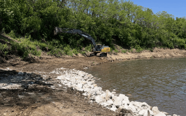 Excavator clearing land by body of water, trees and rocks nearby.