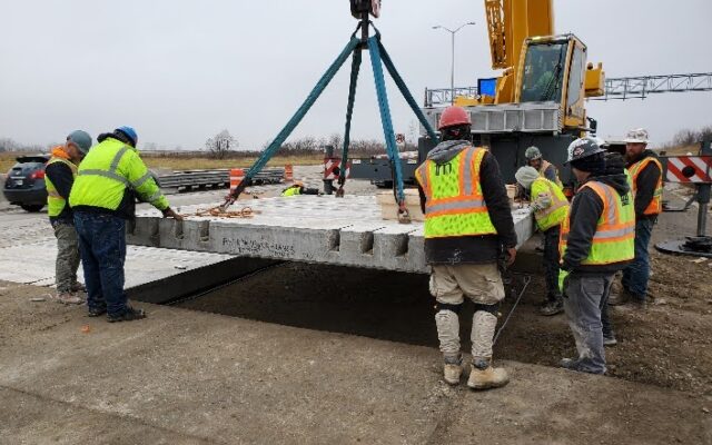 Workers in safety gear guide crane-lifted concrete slab roadside.
