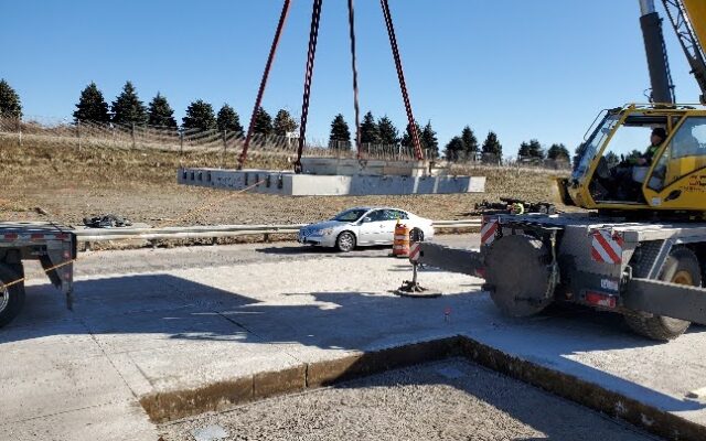 Crane lifts concrete slab near white car and trailer.
