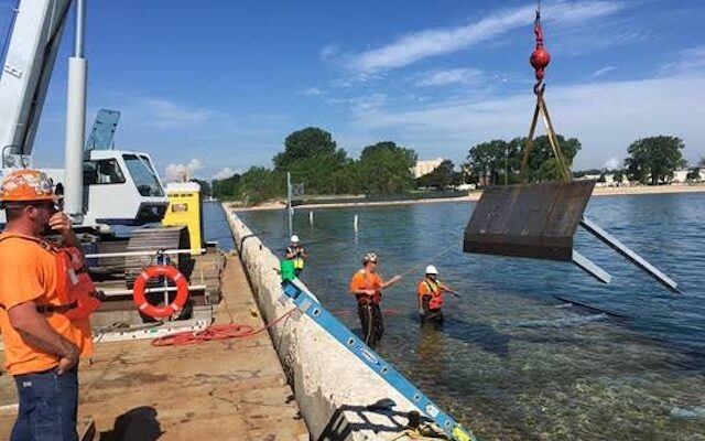 Workers guide crane lifting metal panel from water near pier.