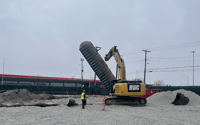 Worker watches excavator lift metal tank at gravel worksite.