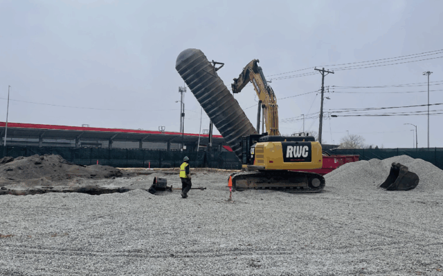 Excavator lifts pipe; two workers stand nearby on gravel.