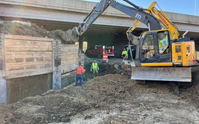 Excavator moves soil by workers under bridge near retaining wall.