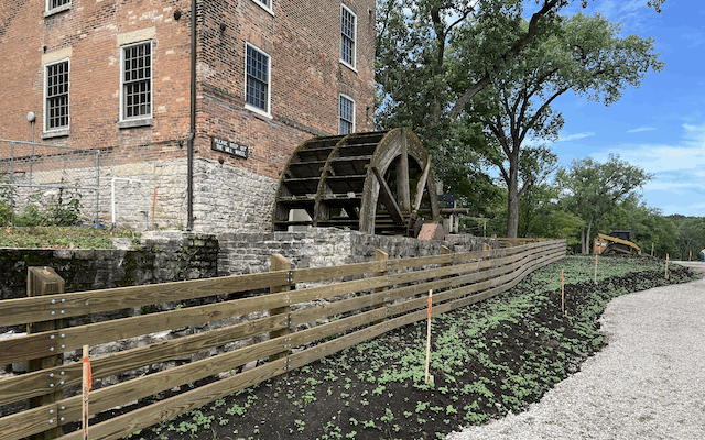 Brick building with water wheel, fence, plants, and gravel path.