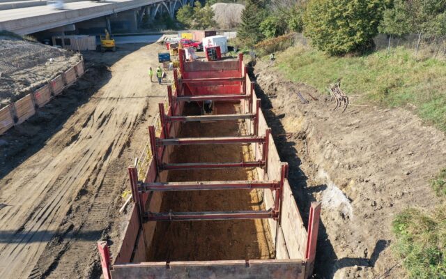 Construction trench near overpass, supported by panels, workers nearby.