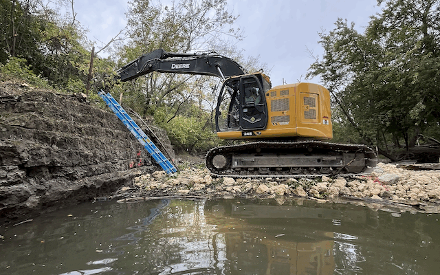 Yellow excavator digs hillside by water, trees and clouds overhead.