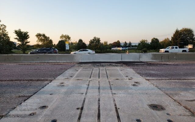 Highway with cars, concrete barrier, trees, and signs visible.