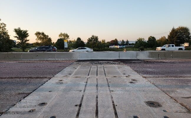 Highway with cars, concrete barrier, trees, and signs visible.
