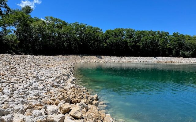 Rocky shore, clear water, dense trees, bright blue sky.
